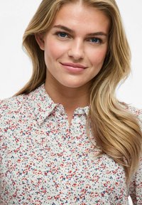 Kwiatowa bluzka na jasnym kremowym tle, w wzór z czerwonymi i niebieskimi plamkami. Zapięcie na guziki z wywiniętym kołnierzykiem.