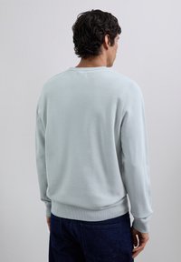 Hombre con cabello rizado oscuro que lleva una camisa de manga larga gris claro y jeans azules oscuros, de espaldas contra un fondo liso claro.