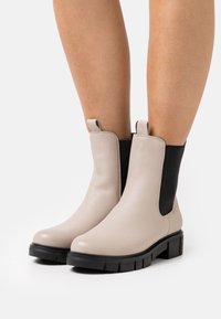 Bottes chelsea en cuir beige avec des panneaux élastiques noirs sur les côtés et une semelle en caoutchouc noir épaisse, dotées de languettes pour un enfilage facile.
