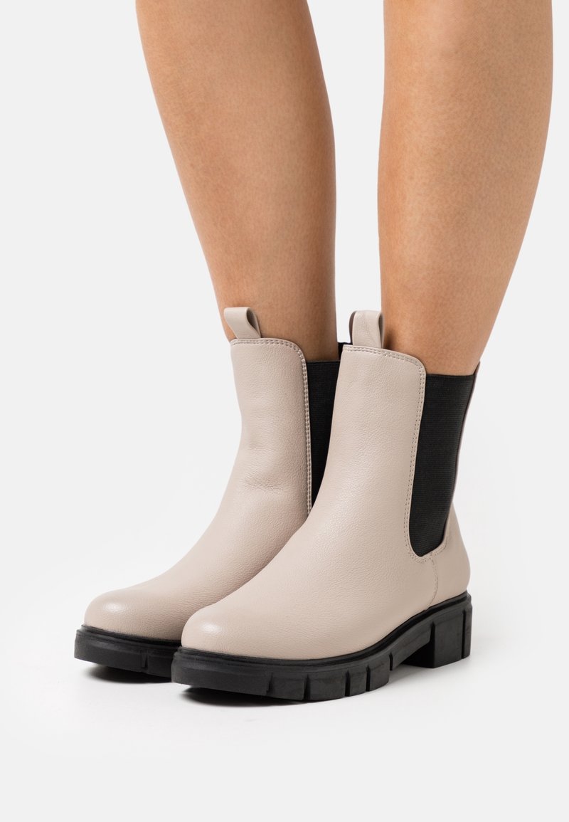 Bottes chelsea en cuir beige avec des panneaux élastiques noirs sur les côtés et une semelle en caoutchouc noir épaisse, dotées de languettes pour un enfilage facile.