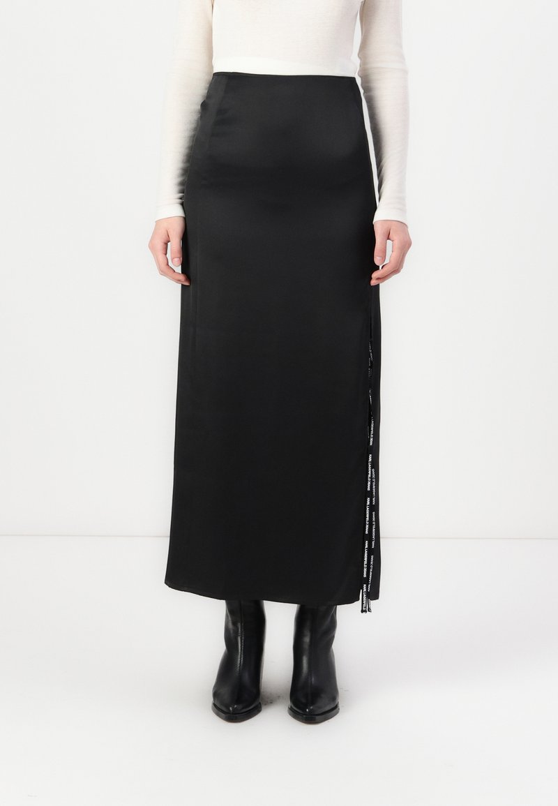 Svarte satin maxi-skjørt med splitt på siden og logo-detalj. Kombinert med tettsittende langermet topp og sorte ankelstøvler. Glatt tekstur.