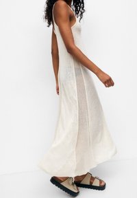 Robe longue beige clair avec fines bretelles, panneaux latéraux en maille, coupe décontractée et ourlet fluide. Associée à des sandales plates de couleur neutre.