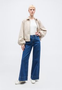 Άτομο με κοντά ξανθά μαλλιά που φοράει μπεζ μπουφάν, λευκό πουκάμισο, μπλε τζιν φαρδιάς γραμμής και λευκά αθλητικά παπούτσια, στέκεται με το χέρι στη μέση.