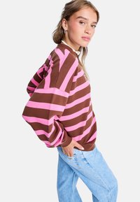 Bruine en roze gestreepte sweatshirt met oversized pasvorm, lange mouwen en geborduurde details. G gedragen met lichtblauwe jeans.