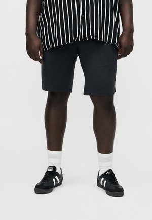 Asmuo, dėvintis juodai baltą dryžuotą marškintę, juodus šortus, baltas kojines ir juodus sportbačius su baltomis juostelėmis, stovintis ant vienspalvio fono.