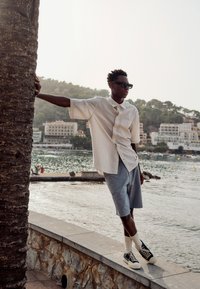 Jeune homme portant des lunettes de soleil, une chemise rayée et un short, appuyé contre un palmier au bord de l'eau avec des bâtiments et des collines en arrière-plan.