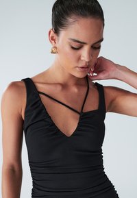 Black sleeveless dress with a V-neck and ruching detail. Features thin crisscross straps and a fitted silhouette. Smooth fabric texture.