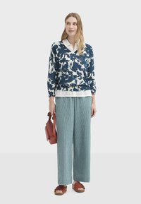 Pull marine à motif floral avec une chemise blanche à col en dessous, associé à un pantalon vert large texturé, un sac à main rouge et des sandales plates.