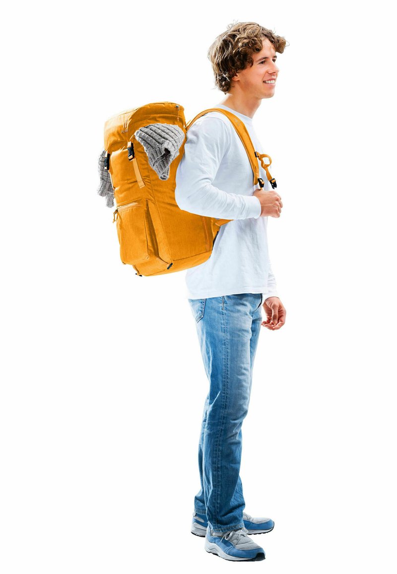 Oranje rugzak met twee zijzakken. Grijze trui die eronderuit steekt. Persoon in een lichte langemouwen shirt en spijkerbroek, staande op een witte achtergrond.