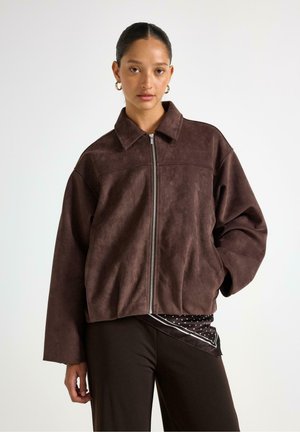 Donna che indossa una giacca marrone scuro con zip e colletto, pantaloni neri e una sciarpa a pois infilata in vita su uno sfondo uniforme.