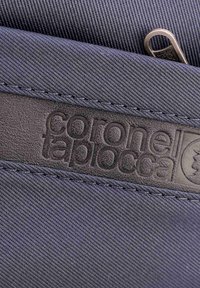 Primer plano de una tela azul oscura con una etiqueta en relieve de "coronel tapiocca" en negro y un tirador de cremallera metálico parcialmente visible.