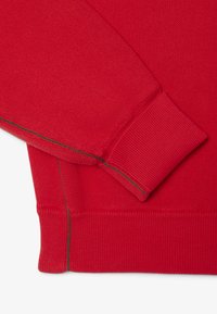 Manche et poignet en tissu rouge texturé avec détail de surpiqûres vertes sur un vêtement rouge assorti, présenté sur fond blanc.