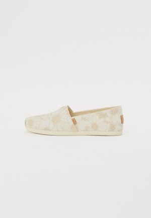 Beige vászon slip-on cipők virágmintával, textúrázott felülettel és kontrasztos barna bőr díszítéssel a sarkán.
