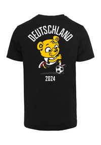 Juodas medvilnės marškinėlis su geltonu meškino grafika, futbolo kamuoliu ir tekstu "DEUTSCHLAND 2024" baltos spalvos. Trumpomis rankovėmis, klasikinio kirpimo.