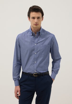 Uomo con camicia a righe blu e bianca con bottoni, pantaloni blu scuro e cintura nera, in piedi con una mano in tasca.