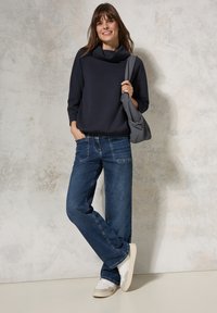 Marinefarbener Cowl-Neck-Pullover mit strukturiertem Muster, kombiniert mit blauen Jeans im Straight-Leg-Schnitt und weißen Sneakers. Trägt eine kleine graue Tasche.