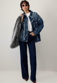 Persona ar nodilušu džinsu jakas, tumšiem džinsiem, baltu krekla, saulesbrillēm, turēt pelēku mākslīgā kažokādas mēteli pār plecu pret vienkāršu fonu.