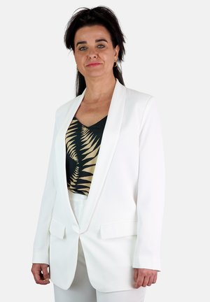 Blazer blanco con solapas de muesca y bolsillos delanteros, combinado con una camiseta sin mangas oscura con un ligero patrón de helechos. Tela suave, corte estructurado.