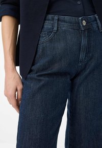 Detailní záběr na osobu v tmavě modrých džínách a černém saku, ukazující pas, kapsy a část levé ruky.