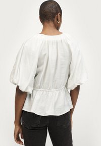 Blouse blanche à manches courtes avec une taille froncée et un design peplum fluide. Le tissu texturé et les larges manches bouffantes ajoutent du volume.