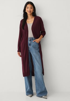 Bordeaux lange cardigan met open voorkant, gebreide manchetten, gedragen over een lichtgrijze top en wijde lichtblauwe jeans, gecombineerd met patroon schoenen.