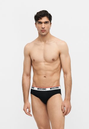 Homme en forme aux cheveux foncés portant un slip noir Moschino avec une ceinture blanche sur un fond clair uni.