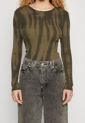 Kobieta ubrana w oliwkowozielony, dopasowany, długi rękaw z ciemnymi pasami tie-dye oraz wysokim stanem wyblakłych czarnych dżinsach, stojąca na jednolitym tle.