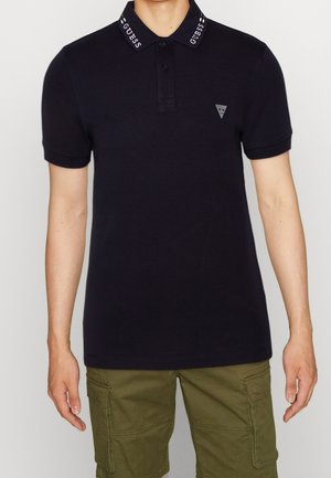 Hombre con camisa polo negra con el texto "GUESS" en el cuello y logo en el pecho, combinado con pantalones cortos cargo de color verde oliva.