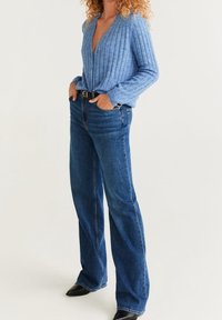 Vaaleansininen ribbineule, jossa on syvä V-pääntie ja pitkät hihat, yhdistettynä korkeavyötäröisiin tummansinisiin farkkuihin ja mustiin nilkkureihin.