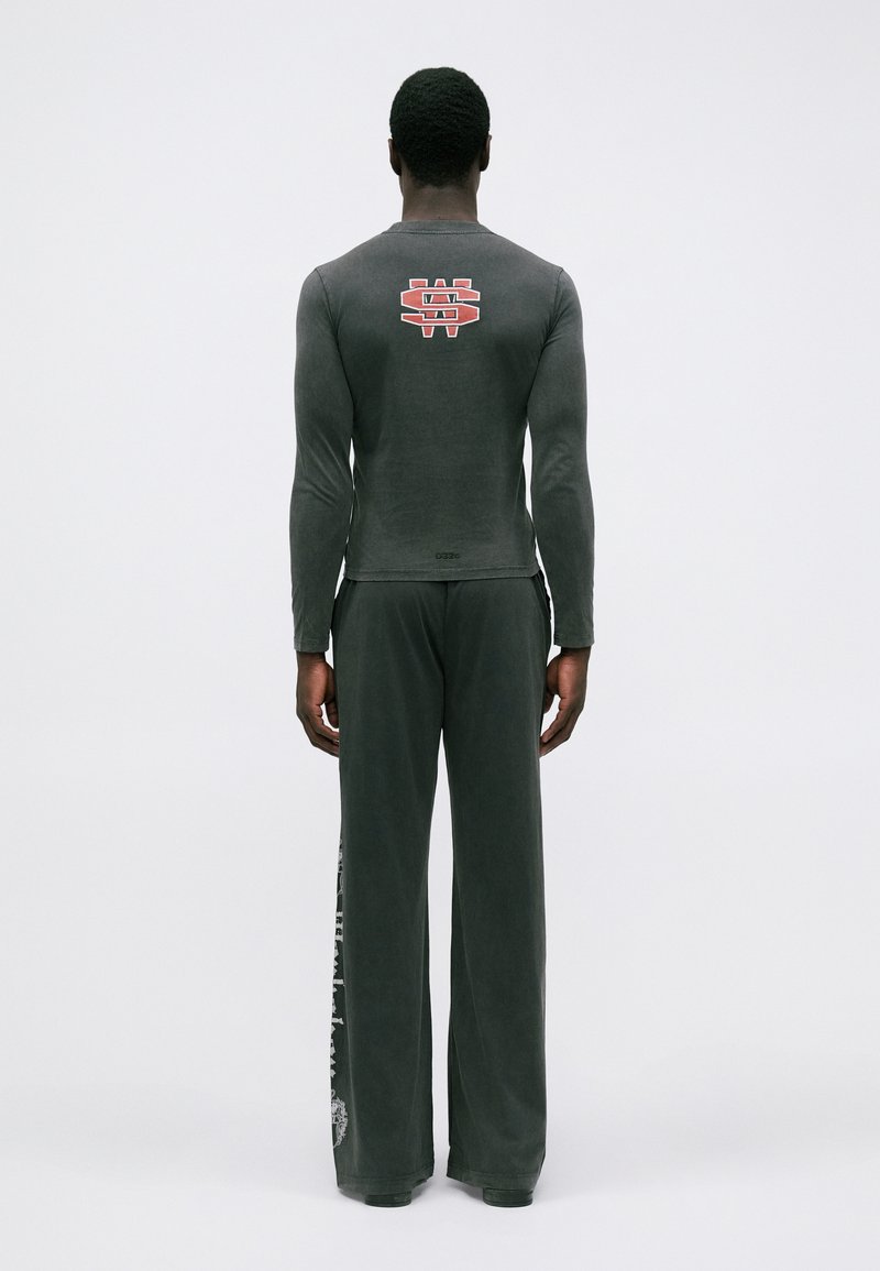 Mand står med ryggen til iført en mørkegrå langærmet skjorte med rødt logo på bagsiden og brede bukser med tryk på venstre ben.