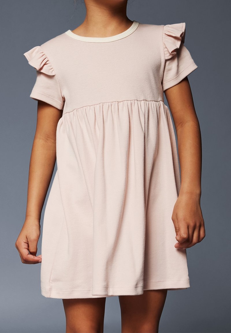 Enfant portant une robe rose clair à manches courtes avec des détails à volants aux épaules sur un fond gris neutre.