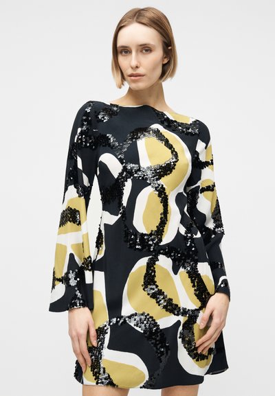Γυναίκα που φοράει μακρυμάνικο μαύρο φόρεμα με αφηρημένα κίτρινα και λευκά σχήματα διακοσμημένα με μαύρες παγιέτες, που στέκεται μπροστά σε απλό φόντο.
