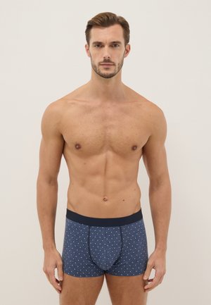 Gespiervde man met kort bruin haar, gekleed in marineblauwe boxershorts met kleine witte stippen, tegen een effen lichte achtergrond.