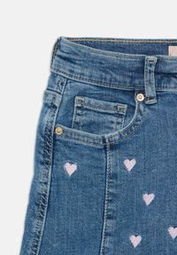 Denim krilo v modri barvi z rožnatimi vezeninami v obliki srca. Ima visok pas, pet žepov in zlate okrasne detajle. Teksture s tkanino.