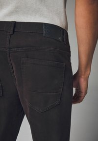 Brown cotton pants feature a textured black waistband patch, two back pockets, and contrast stitching along the seams.