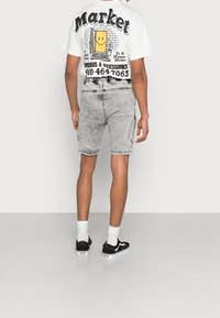 Biała koszulka z grafiką i czarnym napisem oraz uśmiechniętą postacią, w połączeniu z jasnoszarymi szortami dżinsowymi i czarnymi trampkami z białą podeszwą.