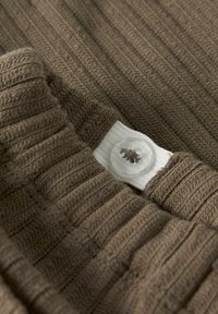 Brunt ribbestrikket stoff med en hvit knapp som har en sirkulær design. Teksturen virker myk med subtile variasjoner i strikkemønsteret.