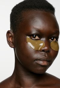 Patch occhi in gel verde oliva su pelle scura e liscia; primo piano del viso con capelli neri corti e labbra naturali su uno sfondo semplice.