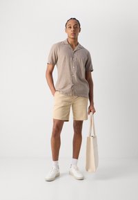 Chemise en lin beige à manches courtes avec des boutons, associée à un short beige clair. Le modèle tient un sac fourre-tout beige, portant des baskets blanches et des chaussettes.