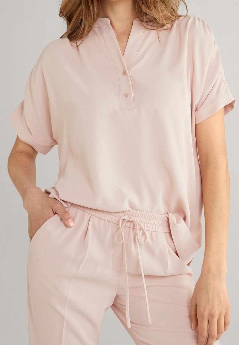 Femme portant un haut ample à manches courtes rose clair avec des boutons et un pantalon à cordon assorti avec des poches, posant avec une main dans une poche.