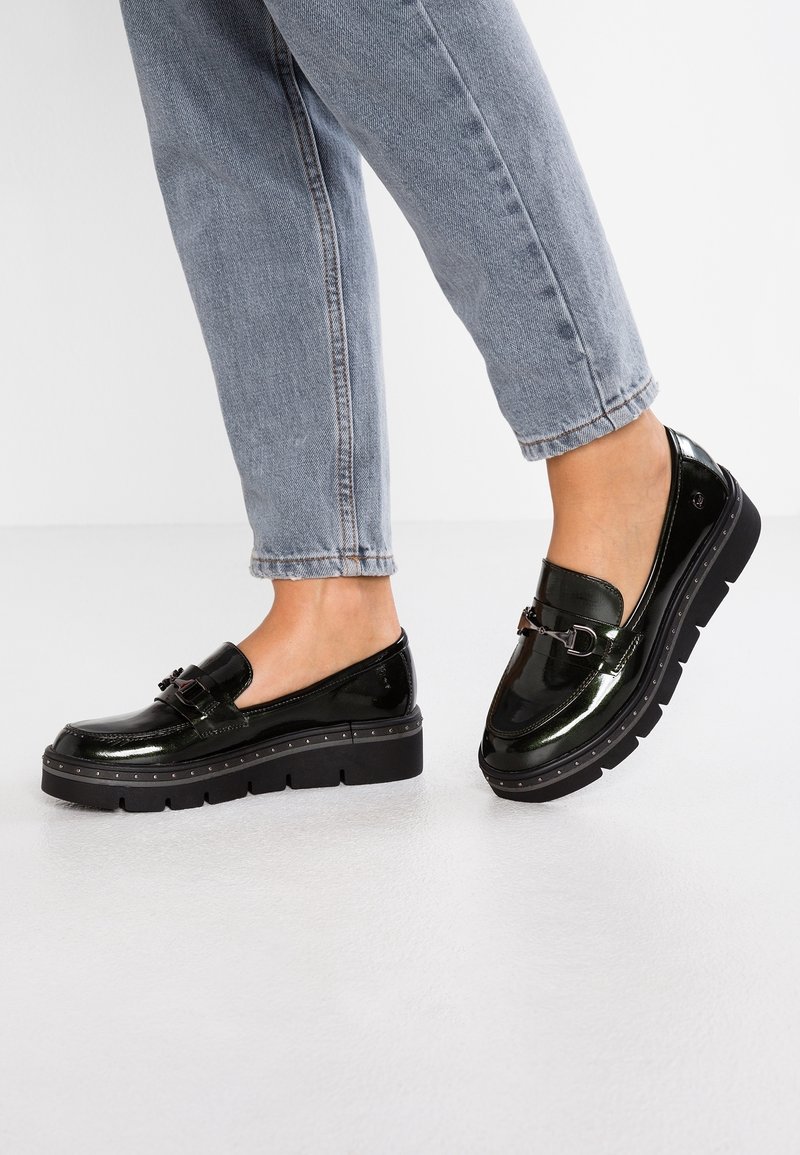 Mocassins à plateforme noirs brillants avec des détails de boucle en métal portés avec un jean bleu clair raccourci sur un fond de sol blanc.