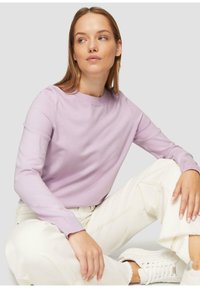 Lila trui met lange mouwen en een ronde hals, gecombineerd met witte omgeslagen jeans en witte sneakers, op een neutrale achtergrond.
