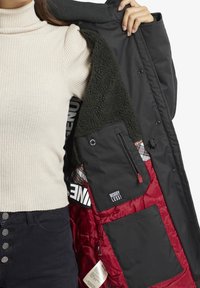 Personne tenant un manteau noir ouvert avec intérieur matelassé rouge, poches intérieures, doublure en polaire, et patch "WORRY LESS !" visible.