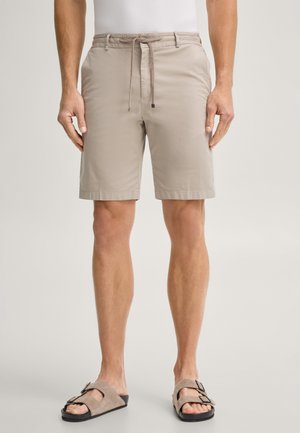 Mann trägt beige knielange Shorts mit Tunnelzug in der Taille und beige Sandalen, steht vor hellem Hintergrund.