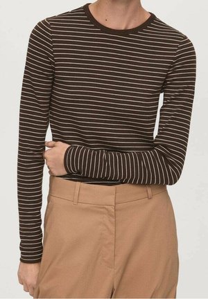 Persoon draagt een bruin shirt met lange mouwen met witte horizontale strepen en beige broek met hoge taille, houdt één arm over de taille.