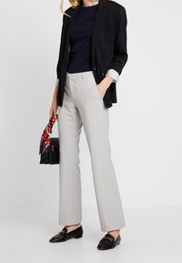 Kvinna i ljusgrå skräddarsydda byxor, svarta loafers, marinblå topp och svart kavaj som håller en svart handväska med en röd blommig scarf.