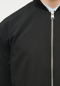 Κοντινό πλάνο ενός ατόμου που φοράει μαύρο μπουφάν με ασημένιο φερμουάρ στο μπροστινό μέρος και μαύρο πλεκτό γιακά πάνω από ανοιχτό γκρι πουκάμισο.