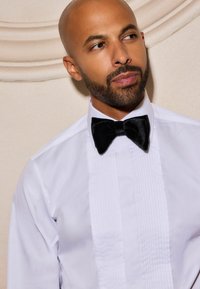 White dress shirt with a pleated front, paired with a black velvet bow tie. Plain background enhances the attire's elegance.