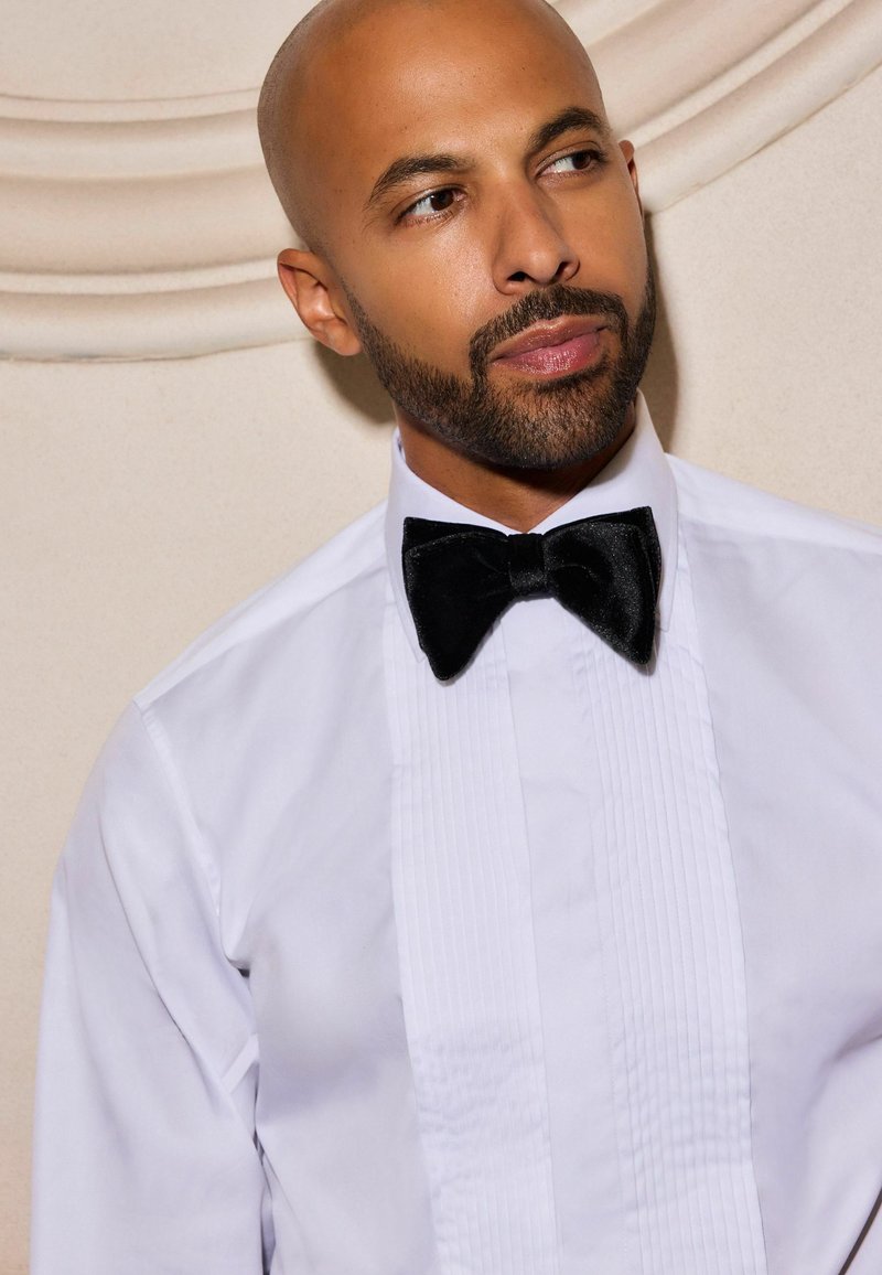 White dress shirt with a pleated front, paired with a black velvet bow tie. Plain background enhances the attire's elegance.