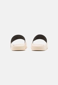 Lys beige sandaler med en teksturert såle og svart, buet stropp. Stroppen har pregete detaljer for grep og stabilitet.